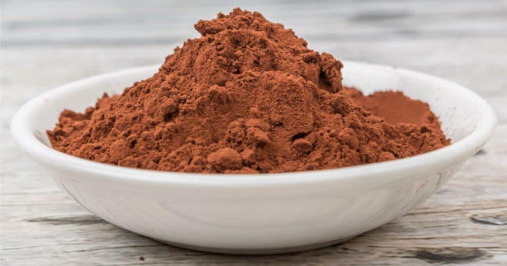 A white bowl filled with a heap of rich, brown cocoa powder sits on a wooden surface, ready for creative baking substitutions.