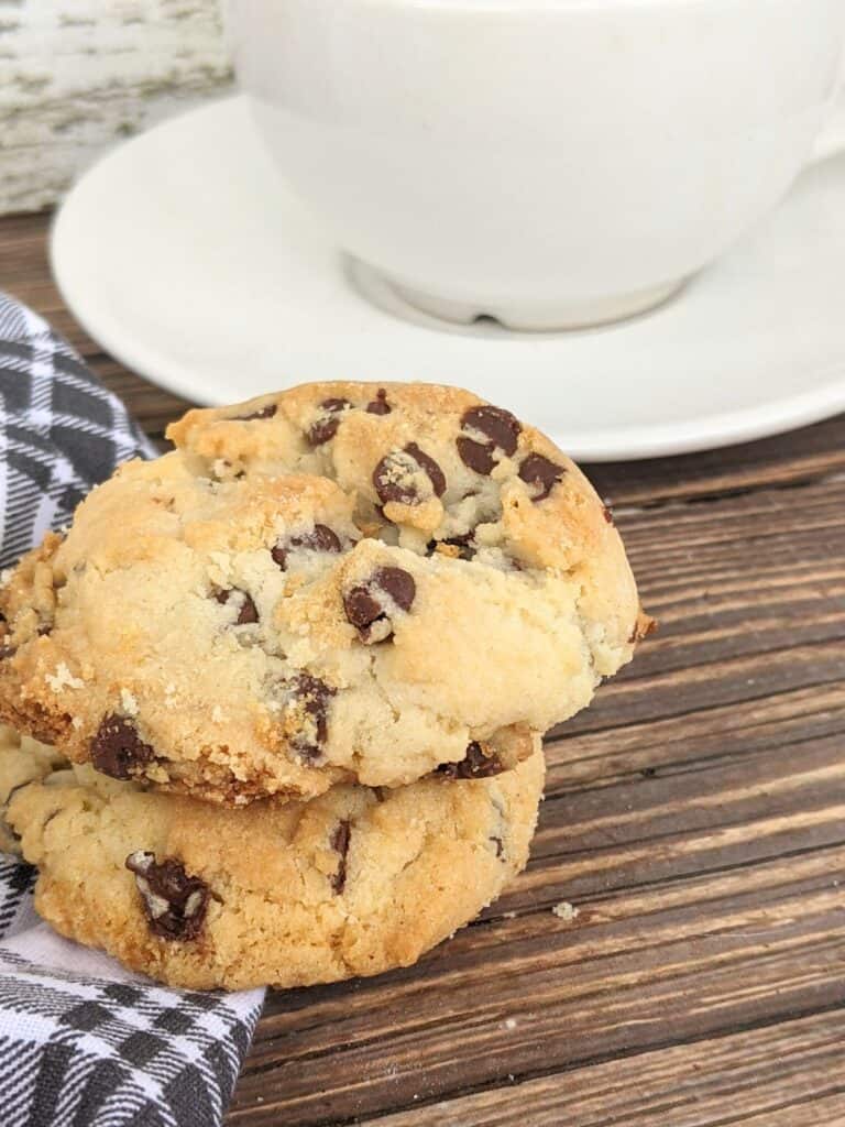 Two chocolate chip cookies stacked on a wooden surface next to a black and white checkered cloth evoke the essence of classic desserts, with a white cup and saucer in the background.
