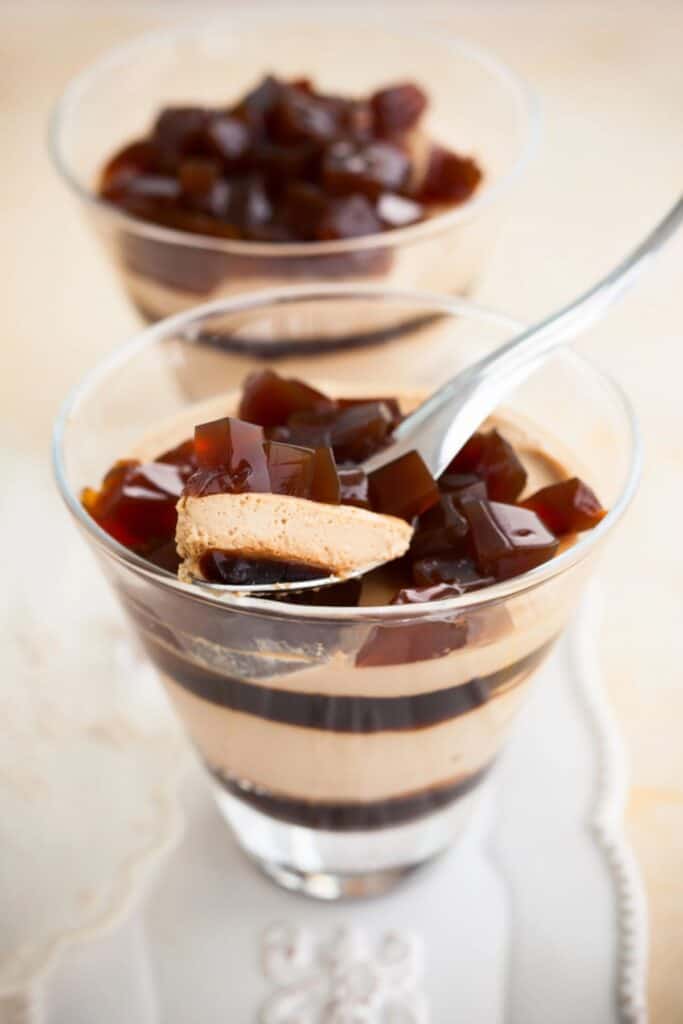 Glass of layered chocolate mousse topped with coffee jelly cubes, with a spoon lifting some coffee-infused jelly. Another similar dessert sits in the background, inviting you to explore this delightful recipe.
