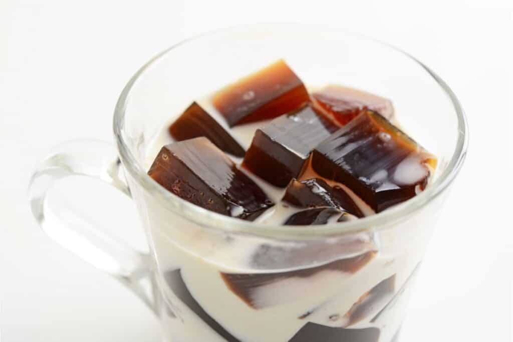 A glass cup brimming with Coffee Jelly cubes and cream rests elegantly on a white background, showcasing a delightful coffee dessert.