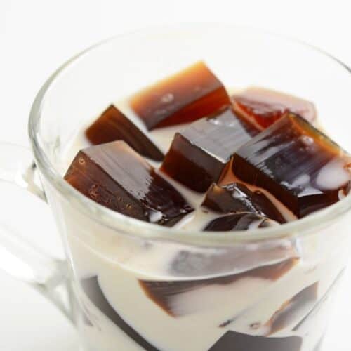 A glass cup brimming with Coffee Jelly cubes and cream rests elegantly on a white background, showcasing a delightful coffee dessert.