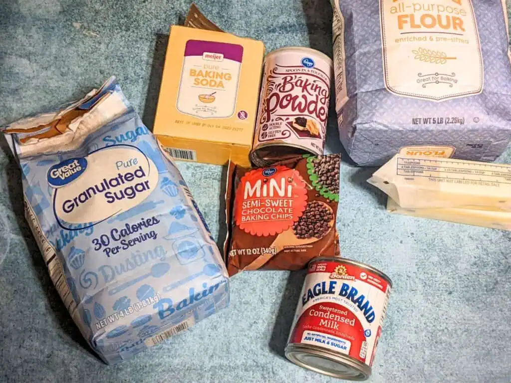 Assorted baking ingredients for your next cookie masterpiece are spread across the surface, including sugar, flour, baking soda, semi-sweet chocolate chips, and a generous helping of butter and condensed milk.