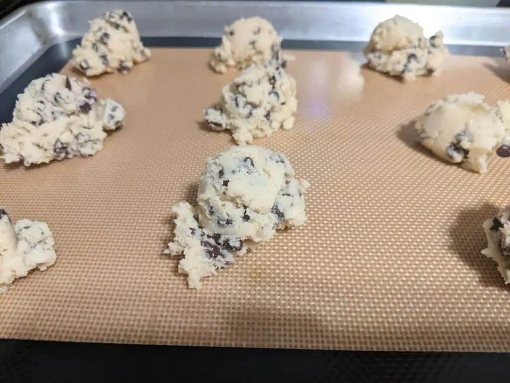 Milk cookies with chocolate chips sit as dough balls on a baking tray lined with a silicone mat, ready for baking.
