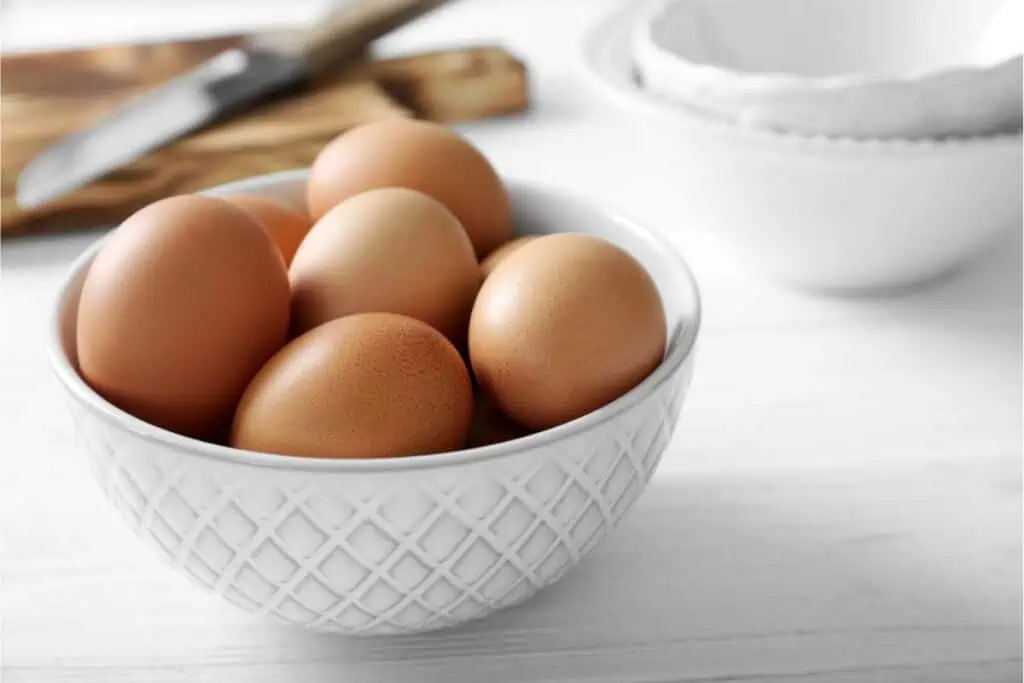 A white ceramic bowl filled with brown eggs sits on a white surface, perfect for all your baking needs. In the background, a knife rests on a wooden cutting board.