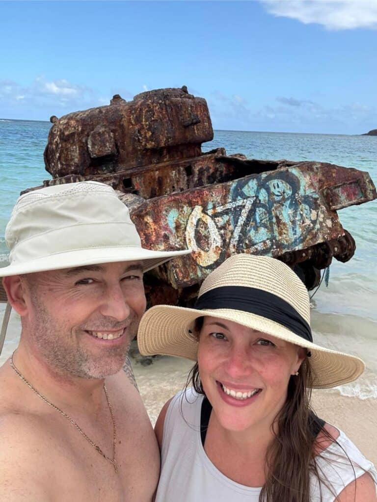 A man and woman wearing hats smile for a selfie on a beach, trying to shake off the post-vacation blues, with a rusty, graffiti-covered tank in the background.