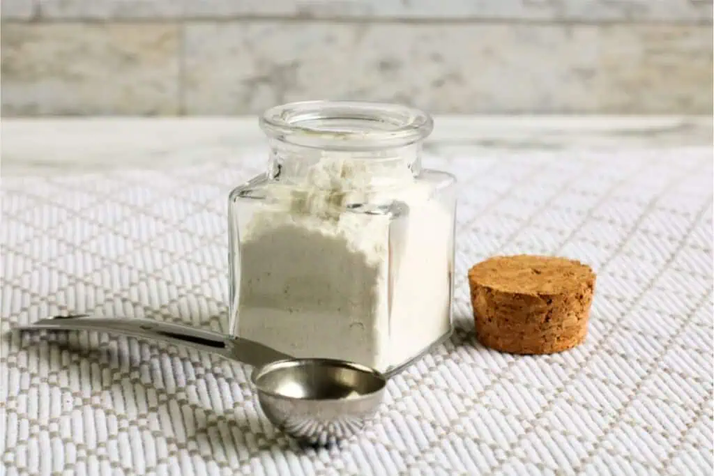 A glass jar filled with white flour, its cork lid set aside, accompanies a metal measuring spoon on a textured white surface—perfect for crafting your own homemade baking powder recipe.
