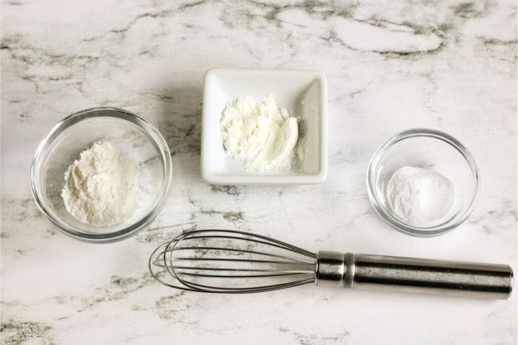 Three bowls containing baking powder, cornstarch, and baking soda sit on a marble surface, waiting to bring a homemade recipe to life. A metal whisk is ready below the bowls.