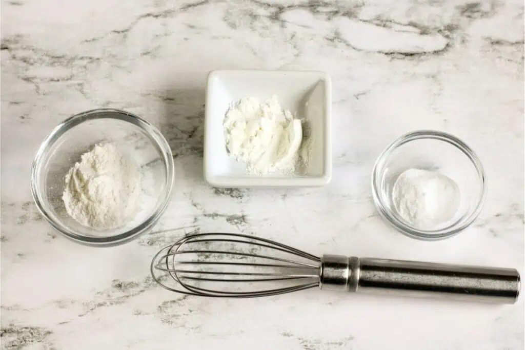 Three bowls containing baking powder, cornstarch, and baking soda sit on a marble surface, waiting to bring a homemade recipe to life. A metal whisk is ready below the bowls.
