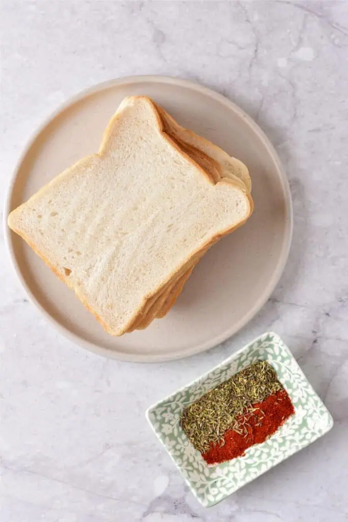 Slices of white bread rest on a round plate beside a small dish containing mixed spices, ready to be transformed into homemade breadcrumbs using your air fryer. The setup is complete on a sleek marble surface.