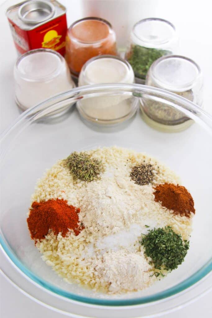 A glass bowl overflows with dry ingredients like homemade breadcrumbs, herbs, spices, and flour. Several jars of spices add a colorful backdrop.