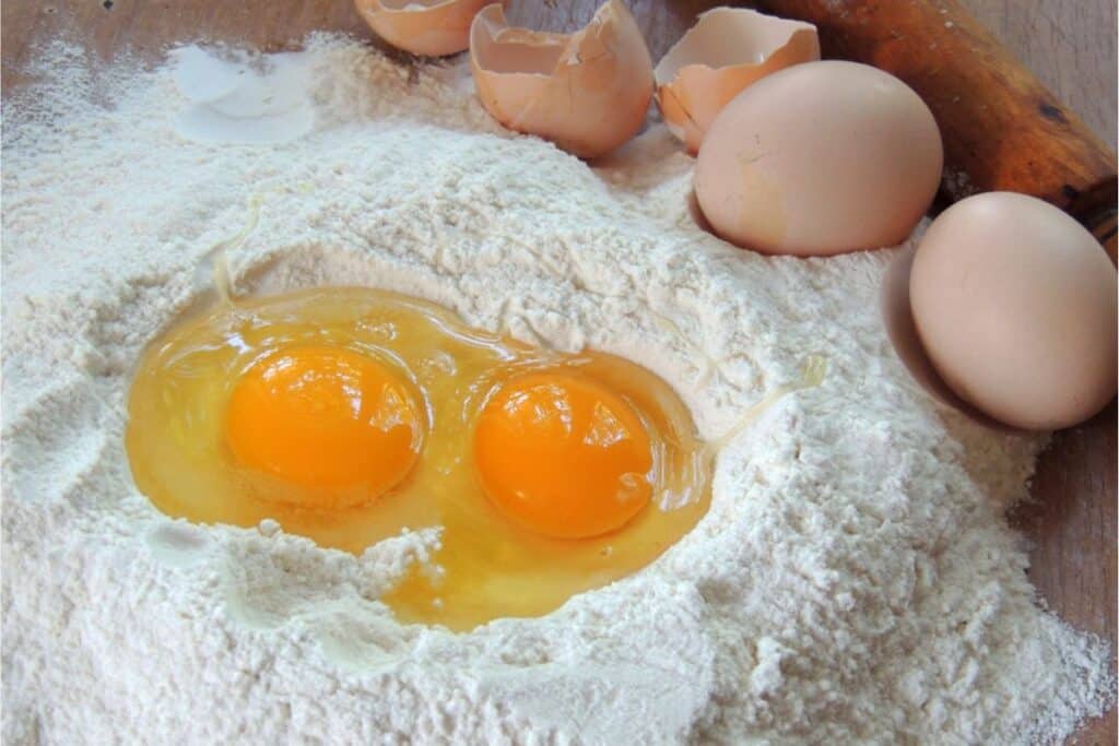 Two cracked eggs with yolks on a pile of flour, surrounded by whole eggs and eggshells, with a rolling pin in the background—just the beginning of a delightful homemade pasta recipe.