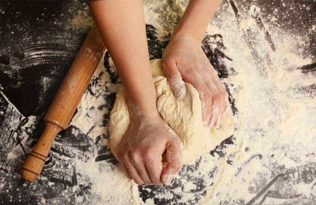 Hands kneading dough on a flour-covered surface, preparing to make homemade pasta, with a wooden rolling pin nearby.