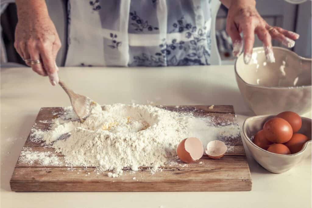 Person preparing homemade pasta dough with flour and eggs on a wooden board, surrounded by a mixing bowl, egg shells, and a bowl of eggs.