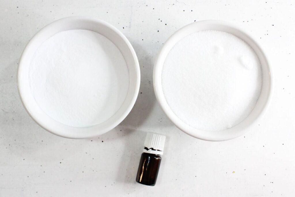 Two white bowls containing white powder, possibly baking soda and sugar, suggest the ingredients for a homemade toilet cleaner, with a small brown bottle placed below them on a white surface.