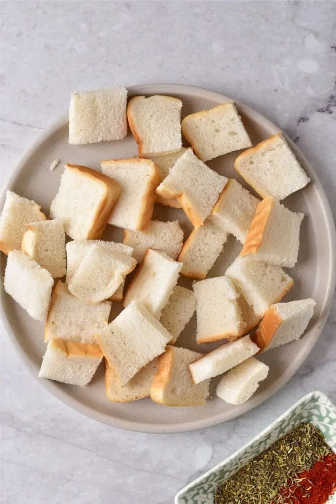 A plate filled with cubes of white bread, perfect for making homemade breadcrumbs, sits on a marble surface. A small dish containing spices is partially visible in the bottom right corner.