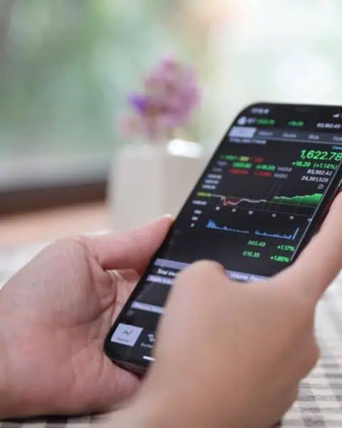 A person holds a smartphone displaying stock market data, with lively graphs and numbers. This snapshot of easy investing takes place against the blurred backdrop of a window and tablecloth, offering beginner investment opportunities at your fingertips.