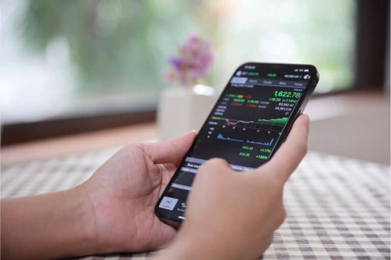 A person holds a smartphone displaying stock market data, with lively graphs and numbers. This snapshot of easy investing takes place against the blurred backdrop of a window and tablecloth, offering beginner investment opportunities at your fingertips.