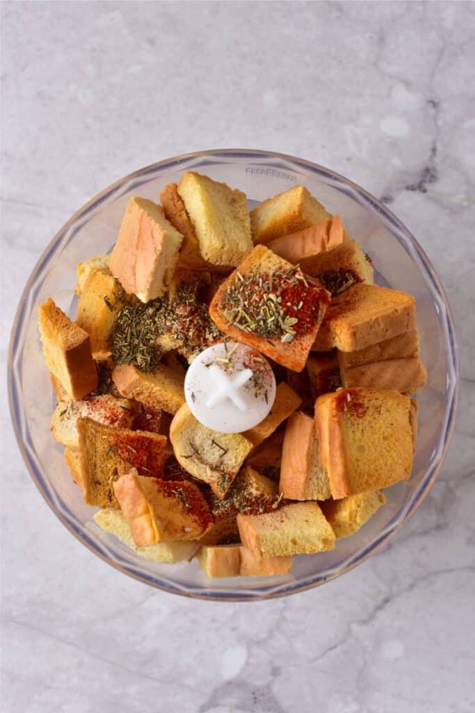 Homemade breadcrumbs and herbs sit in a food processor bowl on a marble surface.