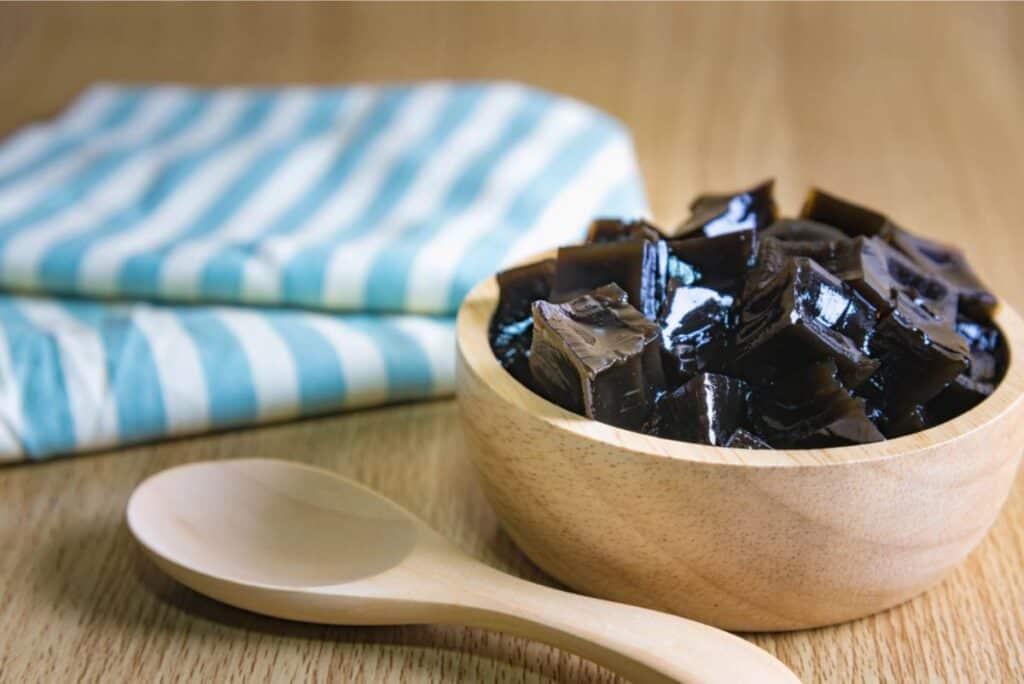 A wooden bowl filled with cubed coffee jelly sits on a wooden surface next to a wooden spoon and a folded blue and white striped cloth.