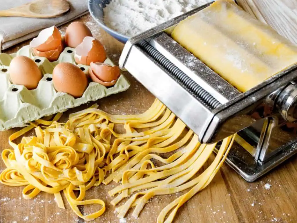 A pasta maker expertly rolls out fresh dough for homemade pasta, surrounded by cracked eggs, egg shells, and a dusting of flour on a rustic wooden surface.
