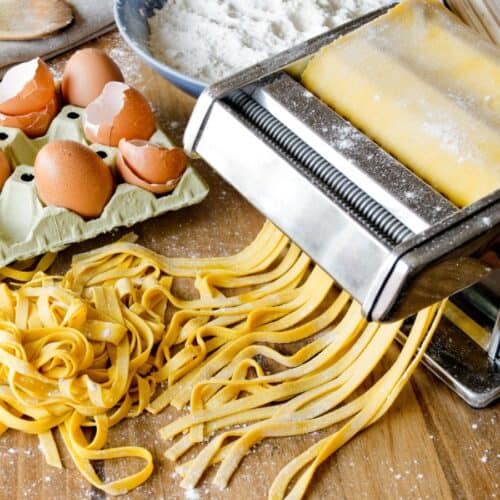 A pasta maker expertly rolls out fresh dough for homemade pasta, surrounded by cracked eggs, egg shells, and a dusting of flour on a rustic wooden surface.