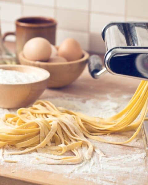 A pasta maker sits elegantly on a wooden surface, surrounded by a bowl of flour and eggs, ready for you to craft your homemade pasta. It’s the perfect setting to dive into your favorite pasta recipe and make fresh noodles from scratch.