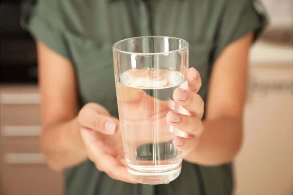 A person wearing a green shirt holds a clear glass filled with water, making sure it's safe to drink.