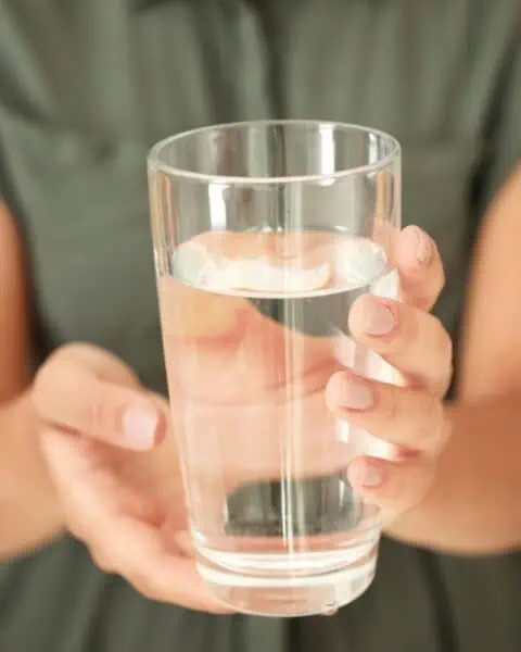 A person wearing a green shirt holds a clear glass filled with water, making sure it's safe to drink.