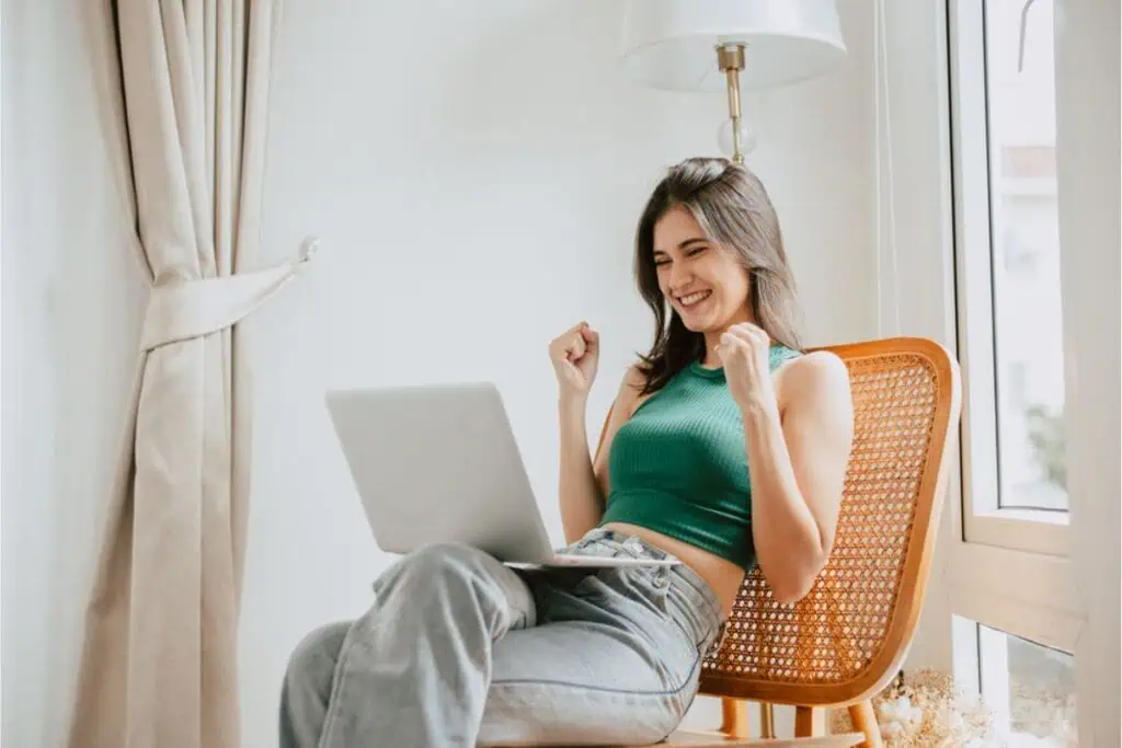 A person sitting in a chair, smiling at a laptop, with fists raised in excitement, celebrates a successful step into beginner investing.