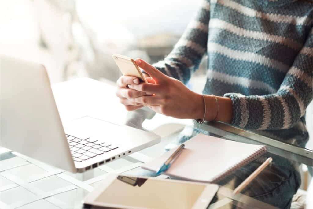 A person in a striped sweater is using a smartphone at a desk, surrounded by a laptop, notebook, pen, and tablet. They appear to be exploring easy ways to invest online, making it accessible even for beginners.