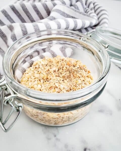 A glass jar filled with chopped oats rests on a marble surface, hinting at a Dry Onion Soup Mix. In the background, a striped cloth adds a touch of rustic charm.