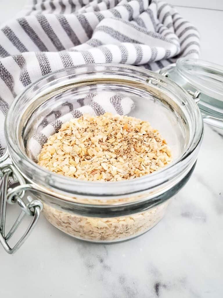 A glass jar filled with chopped oats rests on a marble surface, hinting at a Dry Onion Soup Mix. In the background, a striped cloth adds a touch of rustic charm.