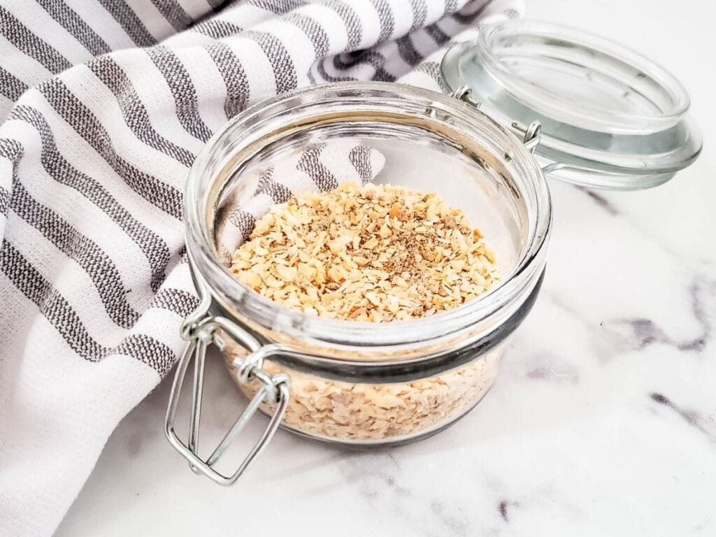 A glass jar of dried minced onions and pepper, reminiscent of a classic onion soup mix, sits on a marble surface. The open lid reveals its aromatic contents, perfectly complemented by the nearby striped cloth.
