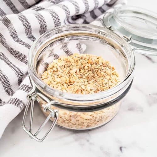 A glass jar of dried minced onions and pepper, reminiscent of a classic onion soup mix, sits on a marble surface. The open lid reveals its aromatic contents, perfectly complemented by the nearby striped cloth.