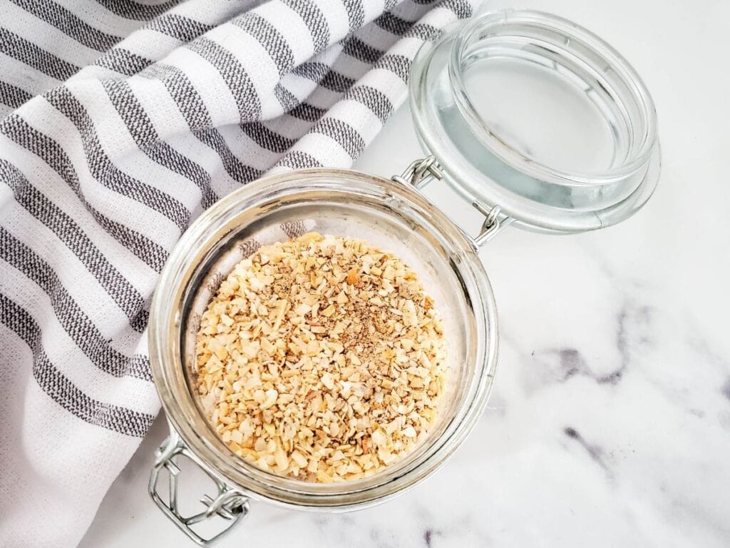 An open glass jar with garlic seasoning sits on a marble surface, next to a striped cloth, ready to elevate any recipe. Perfect for adding depth to soups like your favorite dry onion soup mix.