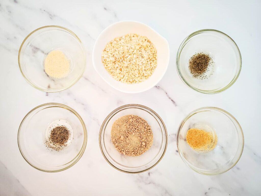 Five small bowls with various spices, including a hint of dry onion soup mix, surround a larger bowl of oats on a white marble surface, setting the stage for an inspiring recipe creation.