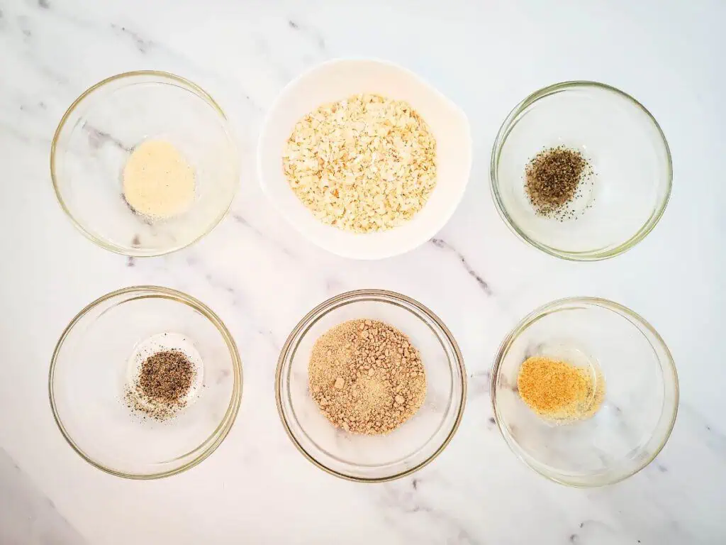 Five small bowls with various spices, including a hint of dry onion soup mix, surround a larger bowl of oats on a white marble surface, setting the stage for an inspiring recipe creation.
