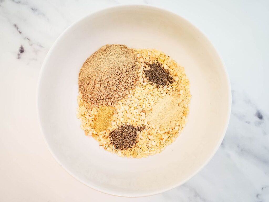 A white bowl on a marble surface holds a vibrant and aromatic blend reminiscent of a dry onion soup mix, featuring four types of spices and herbs that inspire endless recipe possibilities.