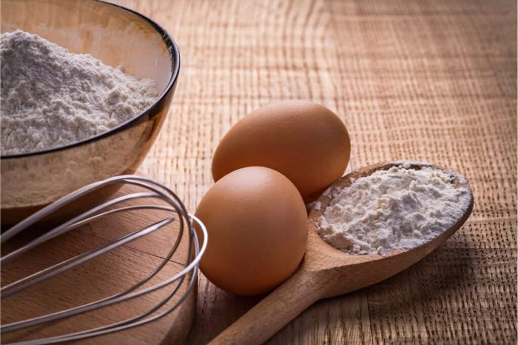 A bowl of flour, two brown eggs, a whisk, and a wooden spoon with flour are ready on a wooden surface for crafting homemade pasta.