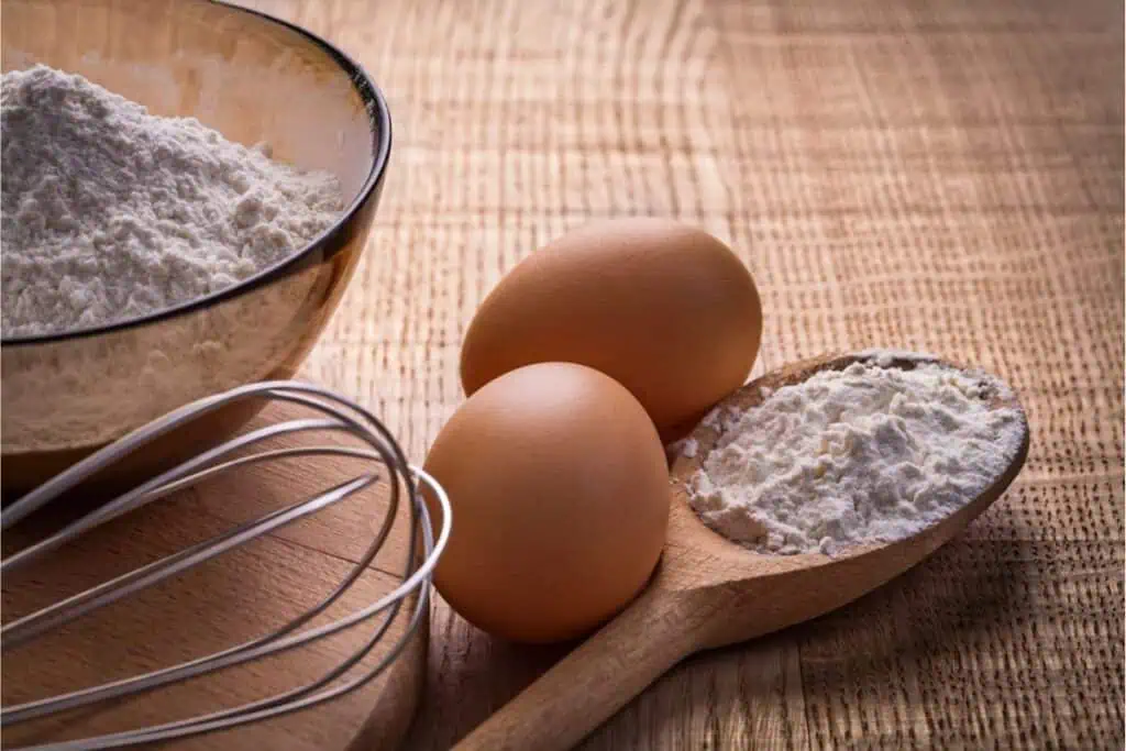 A bowl of flour, two brown eggs, a whisk, and a wooden spoon with flour are ready on a wooden surface for crafting homemade pasta.