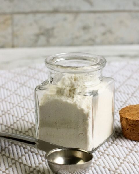 A glass jar filled with white powder, possibly homemade flour, sits on a textured surface. A metal measuring spoon and a cork lid are nearby, hinting at a delicious recipe in the making.