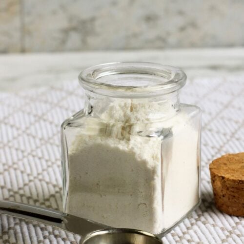 A glass jar filled with white powder, possibly homemade flour, sits on a textured surface. A metal measuring spoon and a cork lid are nearby, hinting at a delicious recipe in the making.