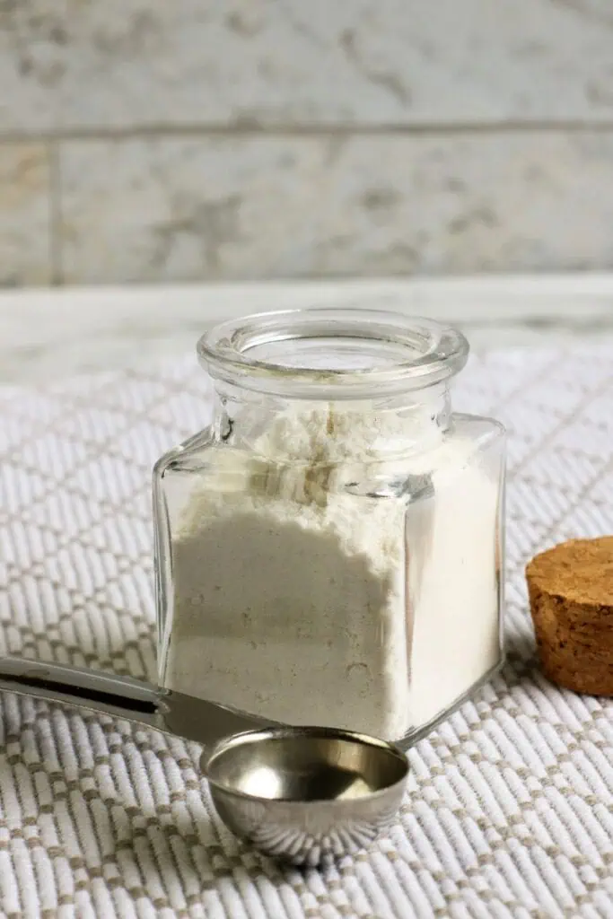 A glass jar filled with white powder, possibly homemade flour, sits on a textured surface. A metal measuring spoon and a cork lid are nearby, hinting at a delicious recipe in the making.