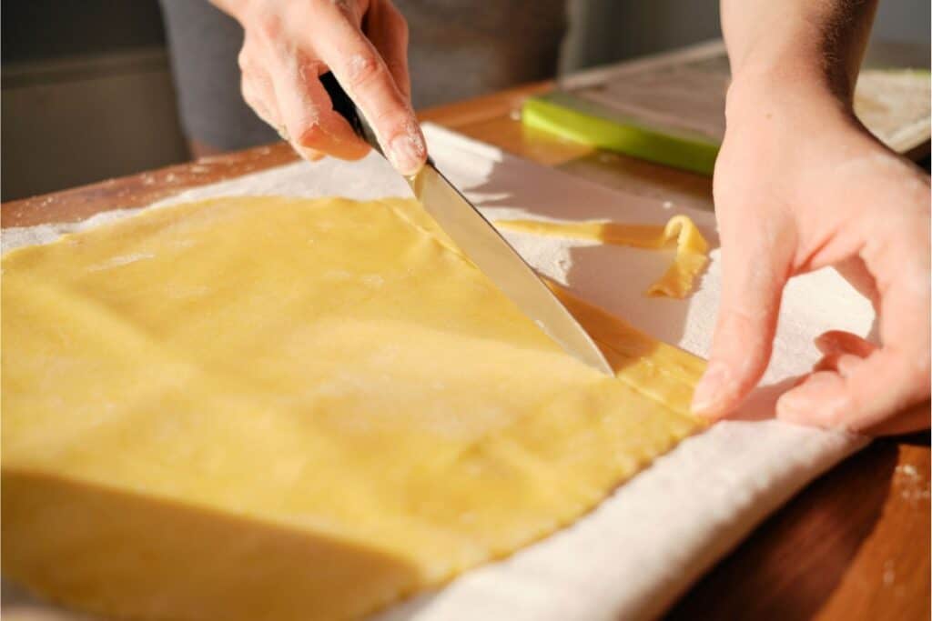 Hands skillfully cutting a sheet of yellow homemade pasta dough with a knife on a floured surface.