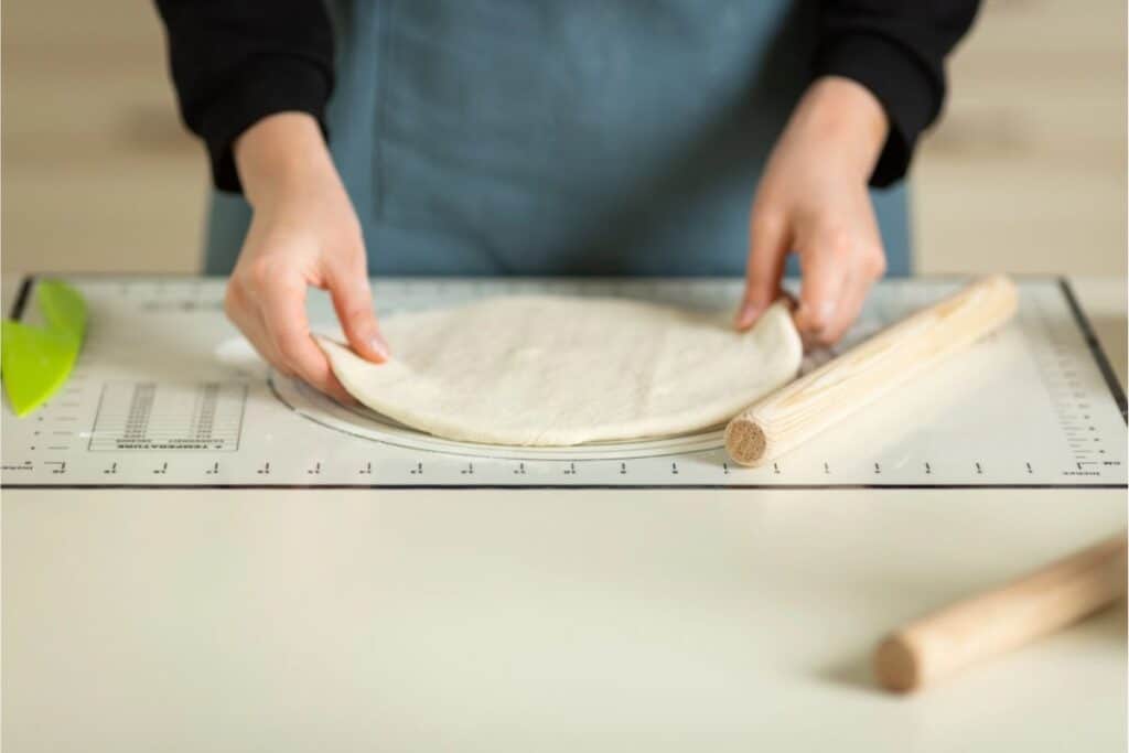 A person wearing an apron shapes dough on a flat surface, perhaps in the midst of exploring a homemade pasta recipe, with a rolling pin resting nearby.