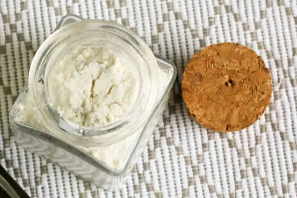 A glass jar filled with a white powder, likely from a homemade baking powder recipe, sits on a textured woven surface. The jar's cork lid is placed beside it.