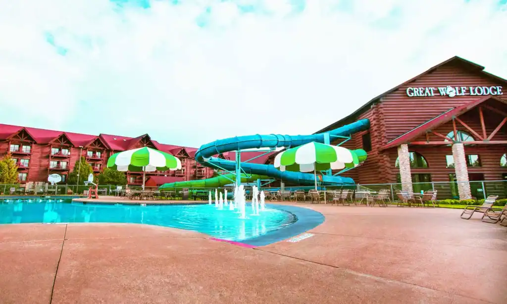 Outdoor pool area at Great Wolf Lodge featuring a waterslide, water fountains, lounge chairs, and red-roofed lodge buildings—experience all this with a Great Wolf Lodge Groupon for extra savings on your stay.