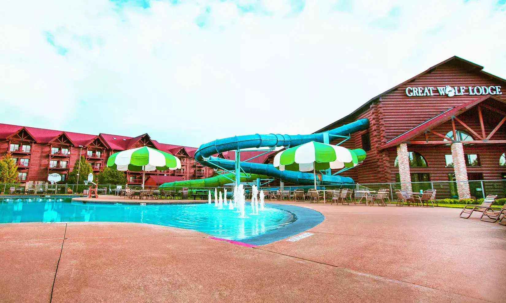 Outdoor pool area at Great Wolf Lodge featuring a waterslide, water fountains, lounge chairs, and red-roofed lodge buildings—experience all this with a Great Wolf Lodge Groupon for extra savings on your stay.