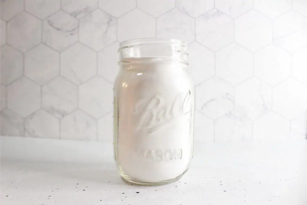 A Glass Mason jar filled with white sugar sits on a white hexagonal tile background, reminiscent of ingredients for an easy homemade toilet bowl cleaner.