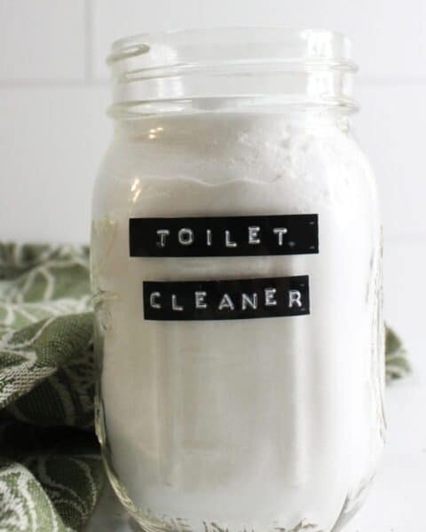 A glass jar labeled "Toilet Cleaner," filled with a white powder mix, sits beside a green and white patterned cloth. This easy homemade toilet bowl cleaner promises to bring sparkle and freshness to your bathroom routine.